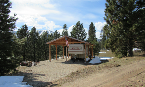A wooden shelter with an RV parked underneath, surrounded by pine trees and patches of snow on the ground.