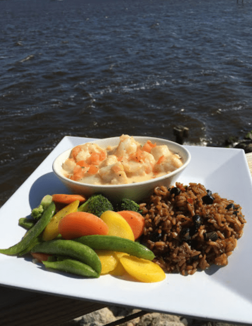 A plate of shrimp and grits, mixed vegetables, and rice, with a scenic view of water in the background.