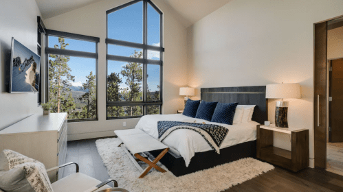 Modern bedroom with large windows, mountain views, a bed with blue pillows, and stylish furniture.
