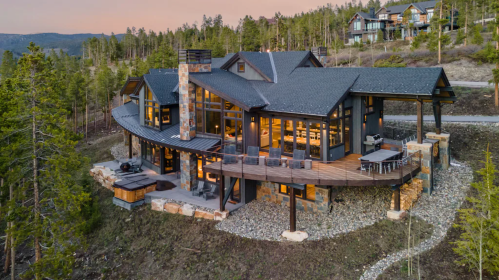 A modern, curved house with large windows, surrounded by trees and mountains, showcasing a scenic sunset view.