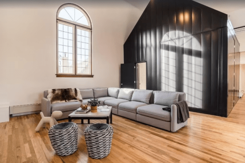 Modern living room with a gray sectional sofa, wooden floors, and a black accent wall. Two woven baskets are in the foreground.