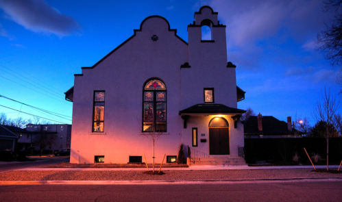 A white church building with arched windows, illuminated at dusk, surrounded by a quiet street and trees.