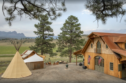 A rustic wooden barn and a teepee surrounded by trees and mountains under a cloudy sky.