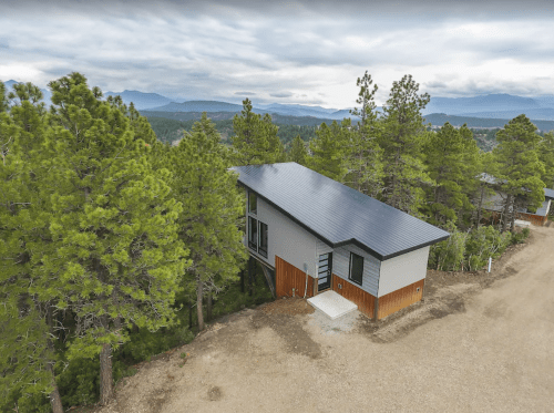 A modern house with a solar panel roof, surrounded by pine trees and mountains in the background.