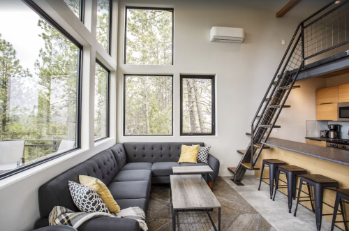 Modern living room with large windows, a sectional sofa, and a staircase leading to an upper level. Forest view outside.