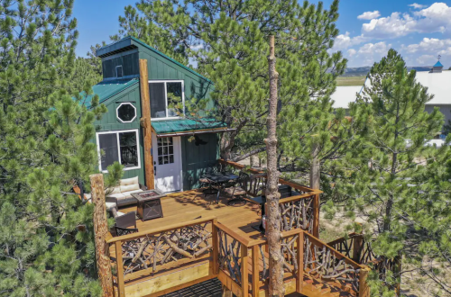 A cozy green cabin surrounded by tall pine trees, featuring a wooden deck with outdoor seating.