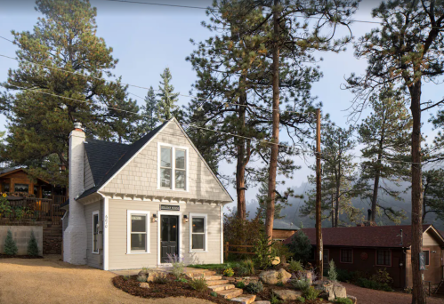 A charming two-story house surrounded by tall pine trees and a gravel pathway, with a cozy atmosphere.