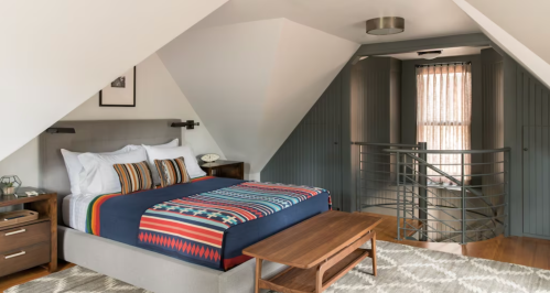 Cozy bedroom with a patterned blanket, wooden furniture, and sloped ceilings, featuring a modern railing and natural light.