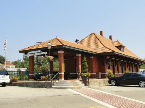 Historic train station with a tiled roof, brick exterior, and surrounding greenery, set against a clear blue sky.