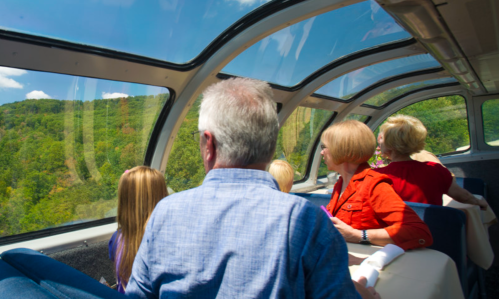 Passengers enjoy scenic views through large windows on a train, surrounded by lush green hills and a clear blue sky.
