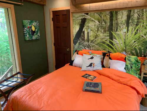 Cozy bedroom with an orange bedspread, tropical decor, and a large window overlooking greenery.