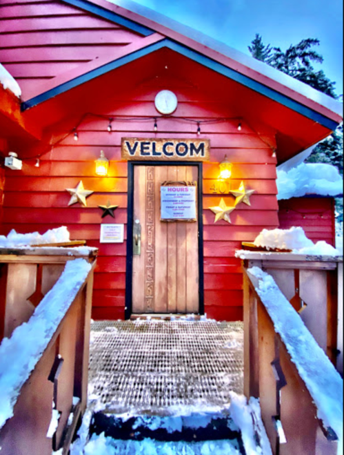 A red building with a wooden door, stars, and a sign reading "VELCOM" surrounded by snow.