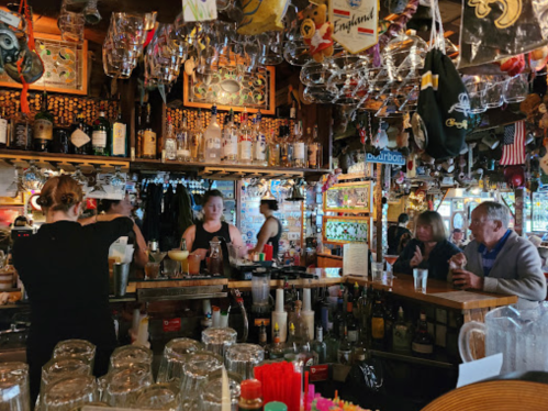 A lively bar scene with bartenders serving drinks, patrons chatting, and colorful decor hanging from the ceiling.