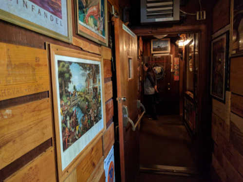A dimly lit hallway with wooden walls and framed artwork, leading to a door where a person stands in the background.