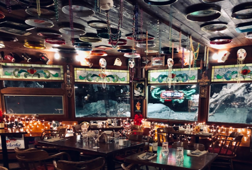 Cozy restaurant interior with colorful plates on the ceiling, string lights, and large windows showing a snowy scene outside.