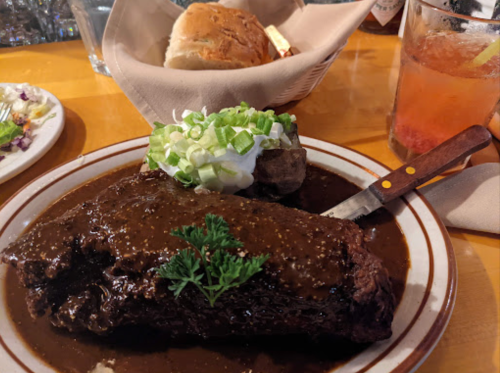 A plate featuring a large piece of beef covered in sauce, a baked potato topped with green onions, and a roll.