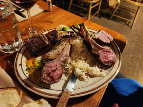 A plate of grilled lamb chops served over rice, garnished with herbs, alongside a glass of red wine.