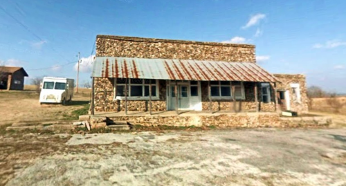 A weathered stone building with a rusty metal roof, set in a rural landscape under a blue sky.