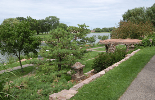 A serene park scene featuring a pond, lush greenery, and decorative stone structures along a walking path.