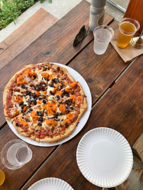 A pizza topped with sausage and cheese on a wooden table, surrounded by empty plates and drinks.