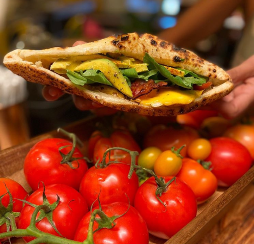 A hand holds a stuffed flatbread with greens, cheese, and vegetables, surrounded by fresh tomatoes on a wooden tray.