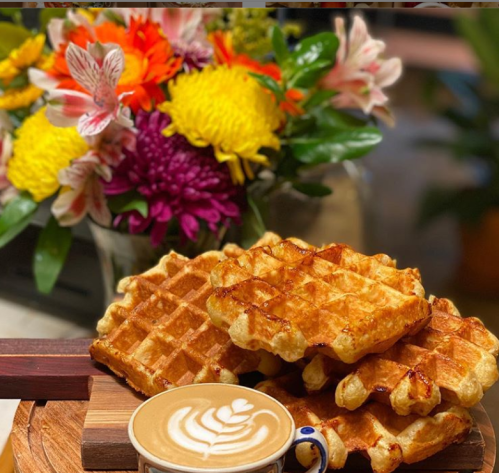 A stack of golden waffles on a wooden board, accompanied by a latte art coffee and a colorful bouquet in the background.