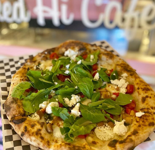 A freshly baked pizza topped with greens, tomatoes, and feta cheese, served on a checkered paper plate.