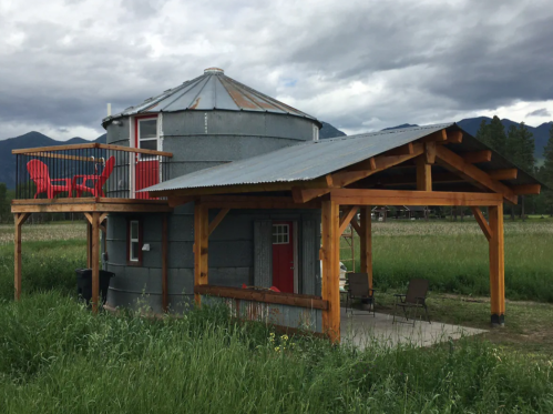 A unique round house with a metal roof, wooden porch, and red accents, surrounded by green grass and mountains in the background.