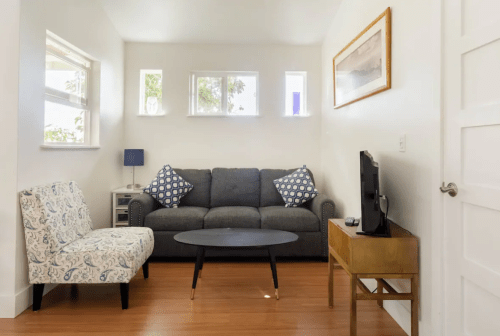 Cozy living room with a gray sofa, patterned chair, coffee table, and TV against a bright, minimalist backdrop.