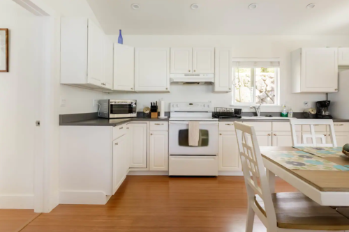Bright, modern kitchen with white cabinets, stainless steel appliances, and a wooden dining table. Natural light from a window.