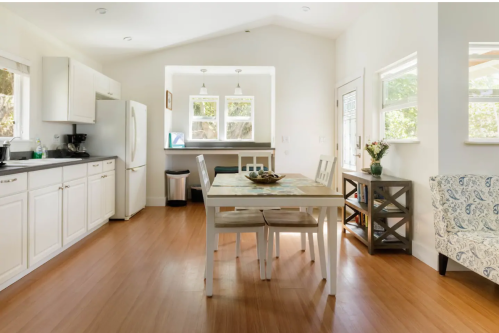 Bright kitchen with white cabinets, a dining table, and a cozy seating area, featuring natural light and wooden floors.
