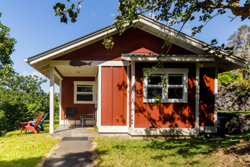 A charming red cottage with a porch, surrounded by greenery and a clear blue sky.