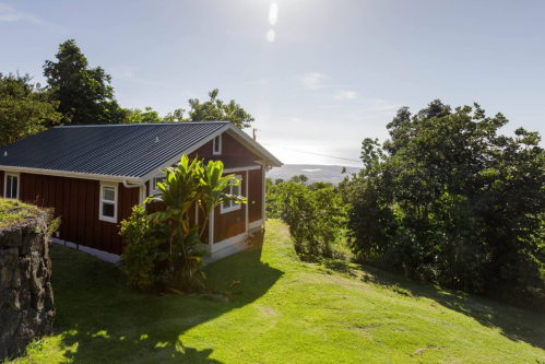 A small red cabin surrounded by greenery, overlooking a scenic landscape under a clear blue sky.