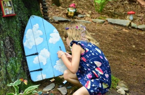 A child in a colorful dress kneels by a whimsical blue door on a tree, surrounded by nature and small decorations.