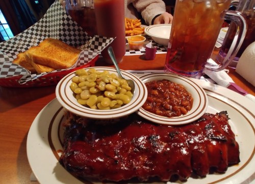 A plate of BBQ ribs with sides of baked beans and lima beans, accompanied by toast and drinks in the background.