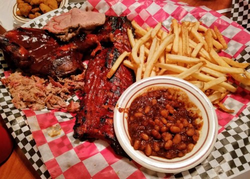 A platter of barbecue ribs, pulled pork, fries, and baked beans on a checkered paper.