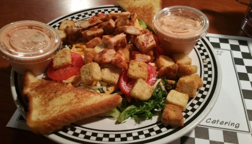 A colorful salad with grilled chicken, tomatoes, croutons, and two cups of dressing, served with toasted bread.