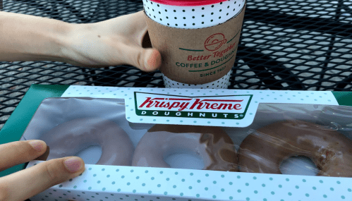 A hand reaches for a box of Krispy Kreme donuts next to a coffee cup on a table.