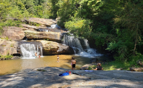 A serene waterfall with people swimming and relaxing by the water, surrounded by lush greenery and rocky terrain.