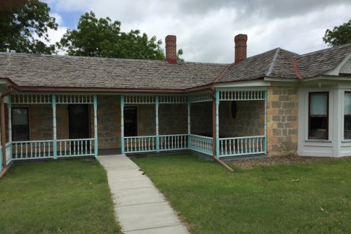 A historic stone house with a porch, green lawn, and cloudy sky in the background.
