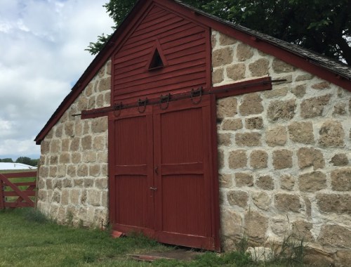 A rustic stone barn with a red wooden door and triangular vent, set against a cloudy sky and green grass.
