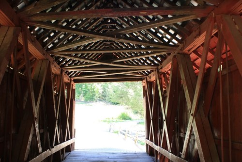 View from inside a wooden covered bridge, showcasing a triangular truss design and light streaming in from the entrance.