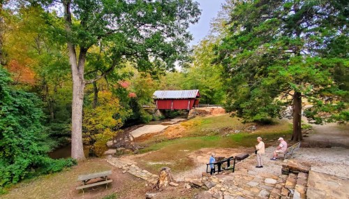 A scenic view of a red covered bridge surrounded by trees, with people sitting and walking nearby.