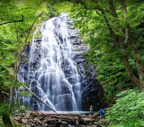 A tall waterfall cascades down rocky cliffs, surrounded by lush green trees, with two people standing at the base.
