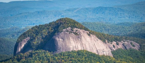 A large, rocky mountain surrounded by lush green forests and rolling hills under a clear blue sky.