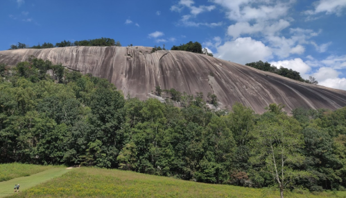 A large, smooth rock face rises above a forested area under a blue sky with scattered clouds.