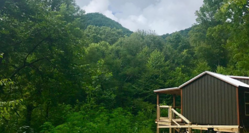 A cabin on a wooden deck surrounded by lush green trees and mountains under a partly cloudy sky.