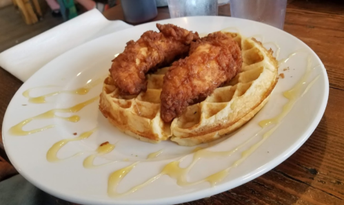 A plate with two crispy chicken tenders on top of a golden waffle, drizzled with syrup.