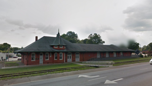 Historic brick train station with a peaked roof, surrounded by greenery and a road in the foreground.