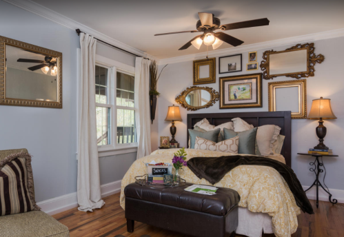 Cozy bedroom with a bed, decorative mirrors, lamps, and a seating area, featuring warm colors and natural light.